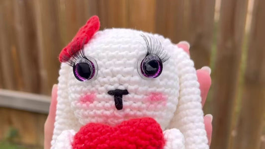 Close-up of a finished white crochet amigurumi bunny with purple safety eyes, lashes, a red bow and a red heart, held up outdoors.