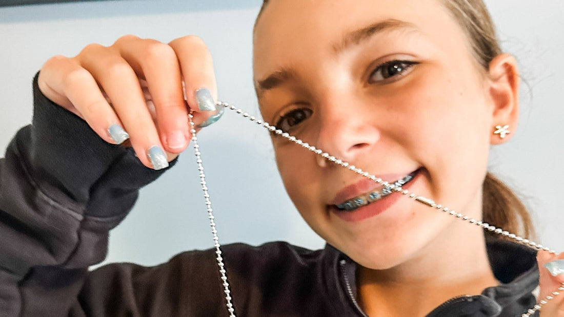 A smiling girl holds up the silver chain of a necklace she made.
