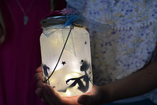 A finished fairy jar glowing in a child's cupped hands, fairy and star silhouettes showing as dark shapes through the frosted tissue paper, with a blue ribbon tied at the neck