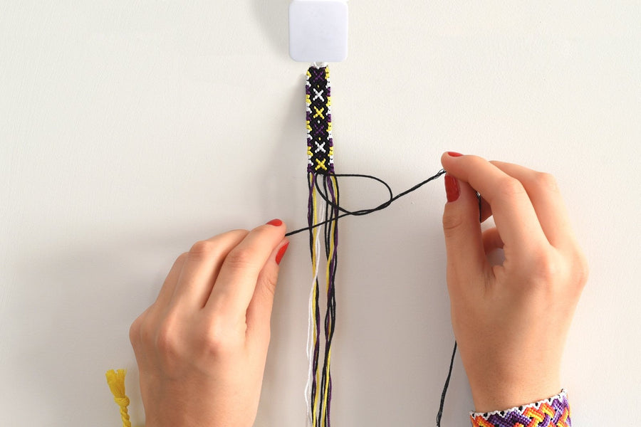 Hands tying a knot on a purple, yellow and white embroidery floss friendship bracelet held in a white braiding clip, with finished bracelets on both wrists.