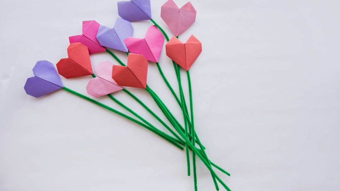 Origami heart flowers folded from red, pink and purple paper on green bendable stems, laid out on a white cloth.