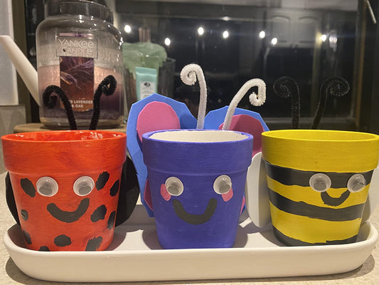 Three finished painted flower pots on a white tray: a red ladybug, a purple bunny and a yellow bumblebee, each with googly eyes and pipe cleaner antennae.