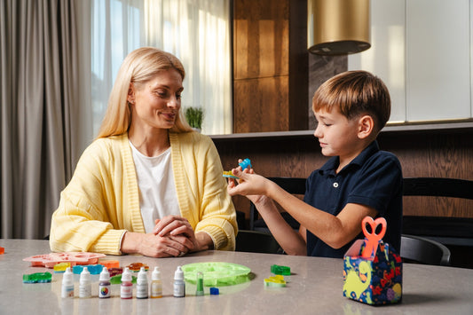 A boy holding a small blue dinosaur soap he has just unmolded, at a kitchen counter with a woman beside him, with silicone molds, colored soap shapes, dropper bottles of soap coloring and a dinosaur-print gift box on the counter