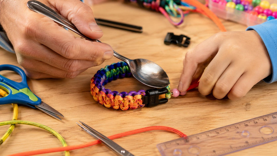 An adult presses the sealed end of a rainbow paracord bracelet flat with the back of a spoon while a child steadies the cord