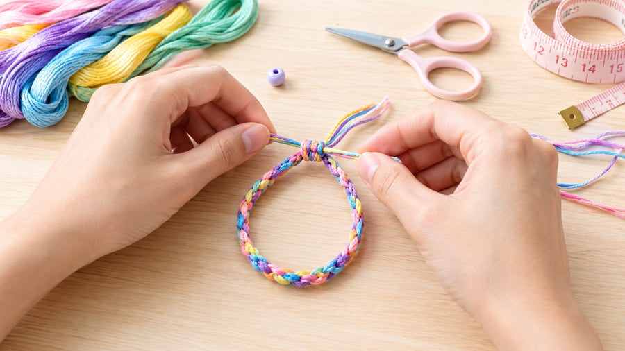 Hands tying the ends of a braided pastel friendship bracelet, with embroidery floss, a bead, scissors, and a measuring tape on the table.