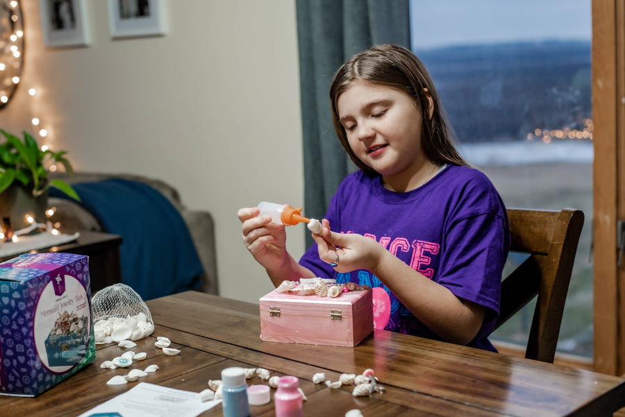 A girl gluing shells onto a pink wooden jewelry box she is decorating at a dining table, with the Mermaid Jewelry Box Kit beside her.