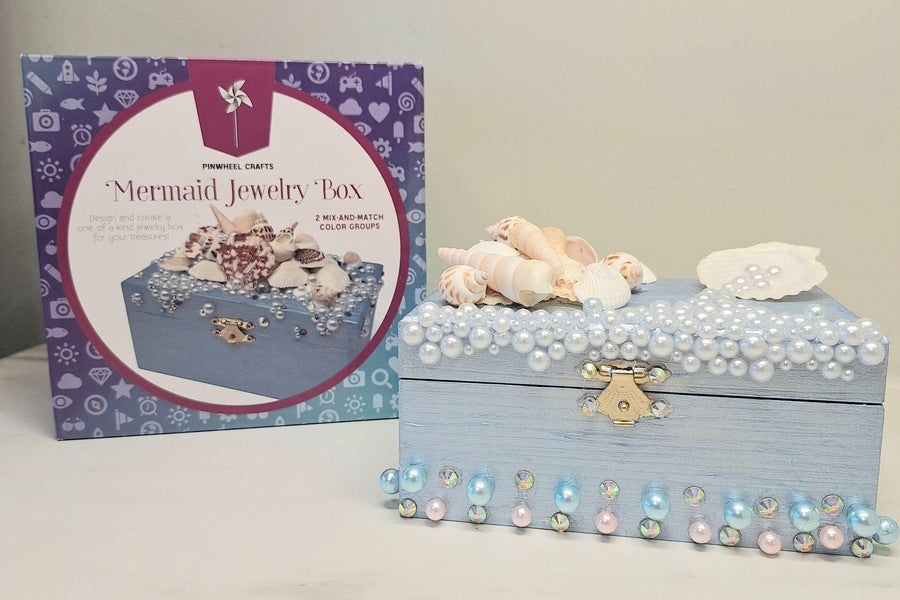 A finished Mermaid Jewelry Box painted pale blue, trimmed with pearls and crystals and topped with sea shells, beside the Pinwheel Crafts Mermaid Jewelry Box kit.