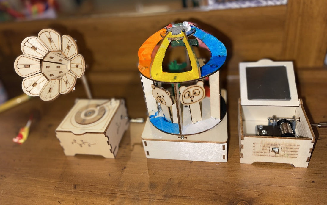 Three finished Music Box Kit builds on a wooden table: a flower gramophone, a carousel painted orange, yellow and blue with bear and panda riders, and an open basic music box showing its metal mechanism.