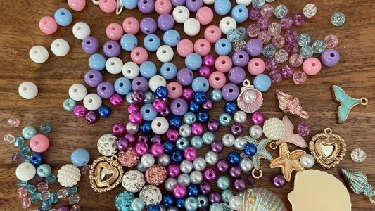 Pastel pink, purple, blue and white beads spread across a wooden table with mermaid tail, seashell, starfish and heart charms from the Mermaid Charm Bracelet Making Kit.
