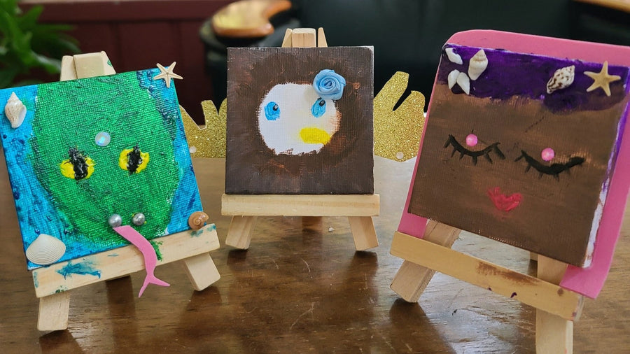 Three finished mini canvases on small wooden easels: a green dragon with yellow eyes, an owl with a blue flower, and a face with purple hair, seashells and a gold star.