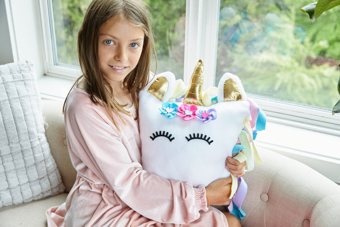 A girl on a window seat hugging her finished white unicorn pillow, its knotted pastel fleece mane hanging down one side