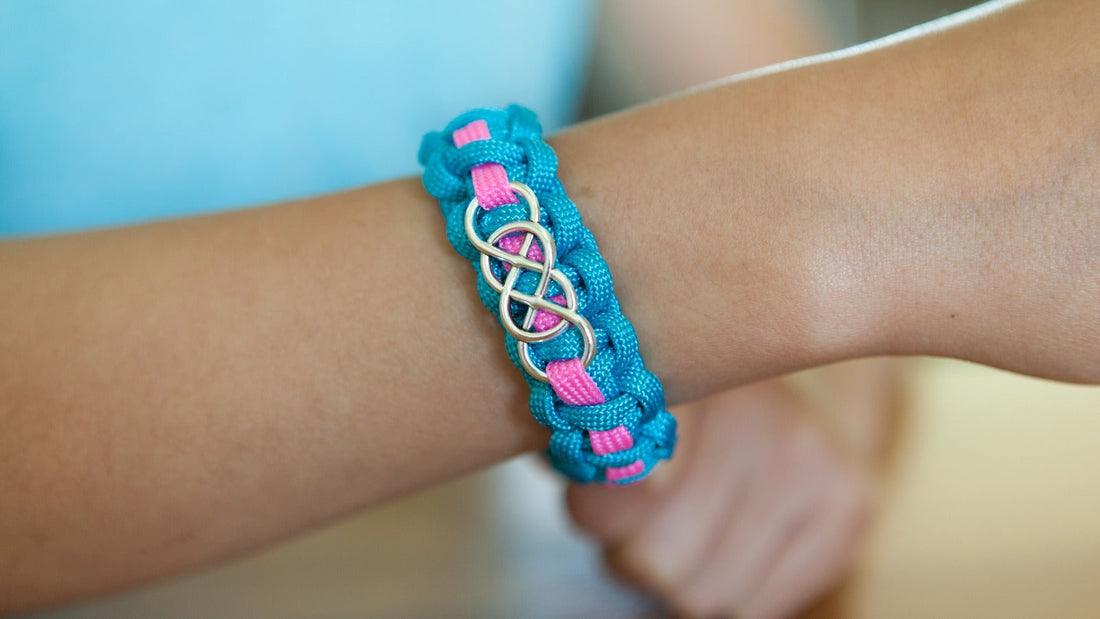 A turquoise paracord bracelet with a pink center stripe and a silver infinity slide charm woven into the knots, worn on a child's wrist.