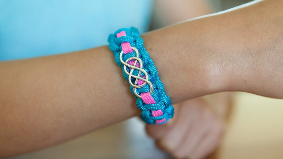 A turquoise paracord bracelet with a pink center stripe and a silver infinity slide charm woven into the knots, worn on a child's wrist.