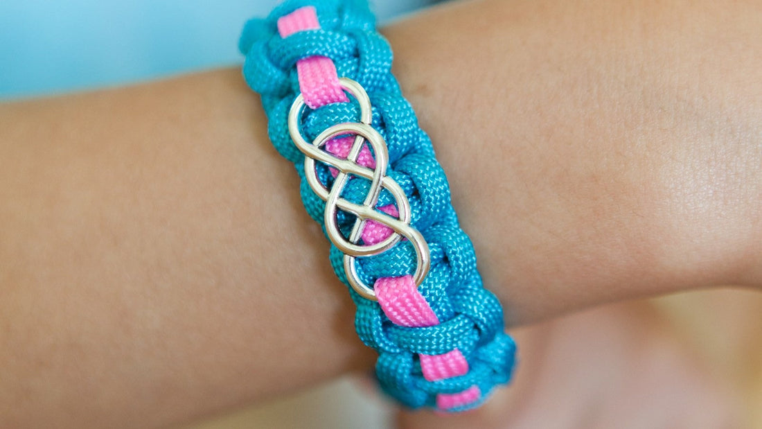 A finished turquoise paracord bracelet with a pink accent stripe and a silver infinity charm on a child's wrist.