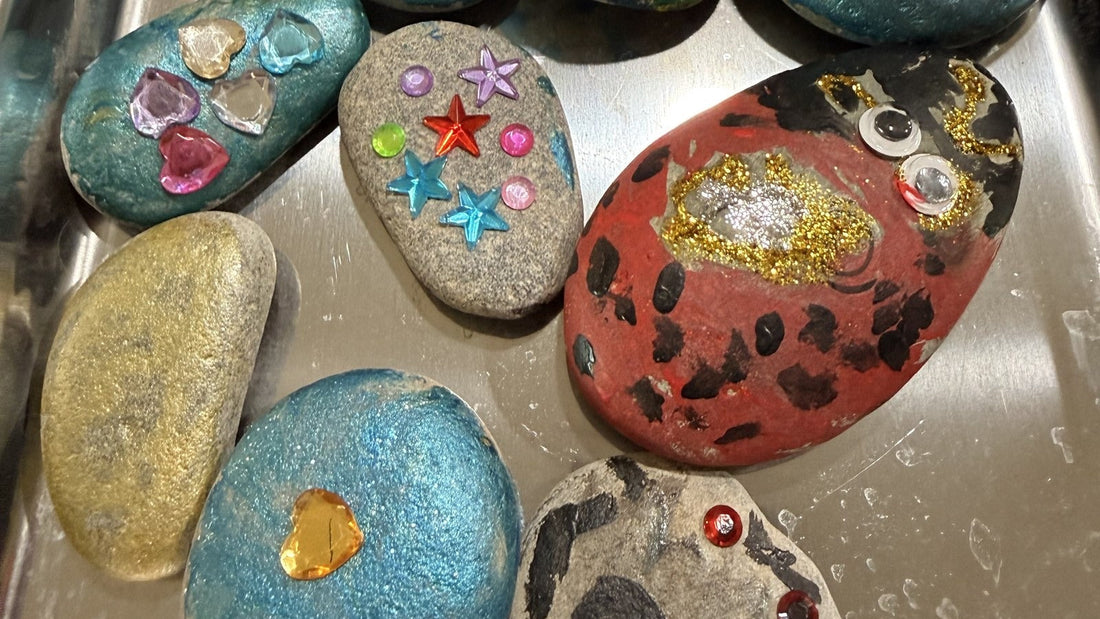 A metal tray of finished painted rocks in metallic blue, gold and red, decorated with gems, star stickers, googly eyes and glitter glue.