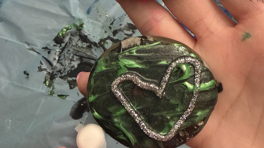 A rock painted shiny green with a silver glitter heart, held in an open hand over a paint-spattered table cover.