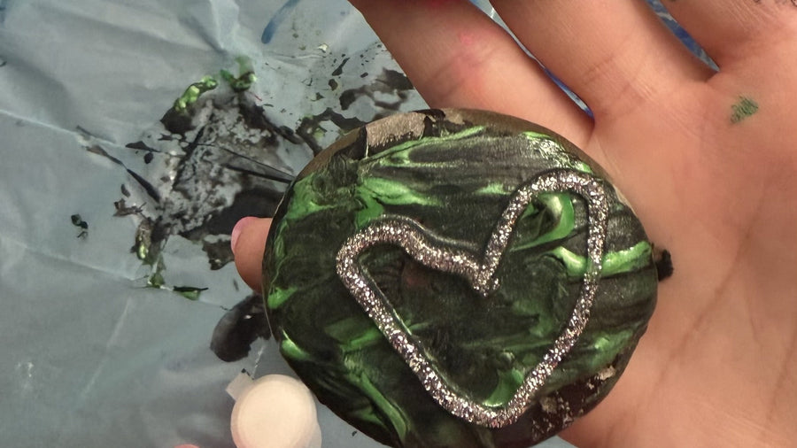 A rock painted shiny green with a silver glitter heart, held in an open hand over a paint-spattered table cover.