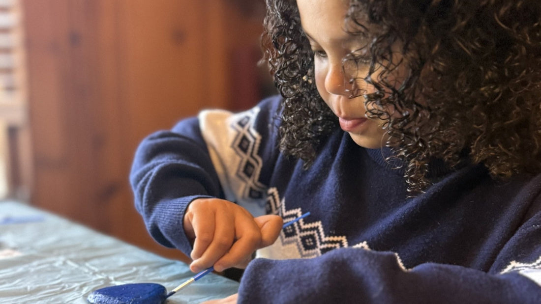 A child in a navy sweater paints a smooth rock blue at a table covered with a plastic cloth.
