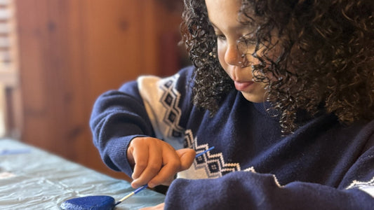 A child in a navy sweater paints a smooth rock blue at a table covered with a plastic cloth.