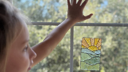 A child reaches up to a sunny window where a sunset suncatcher hangs by its chain from the window's sash bar.