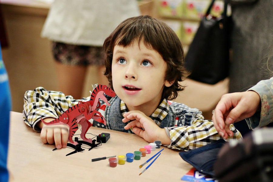 A boy at a table with his finished red robotic T-rex, its wired controller, paint pots and brushes beside it