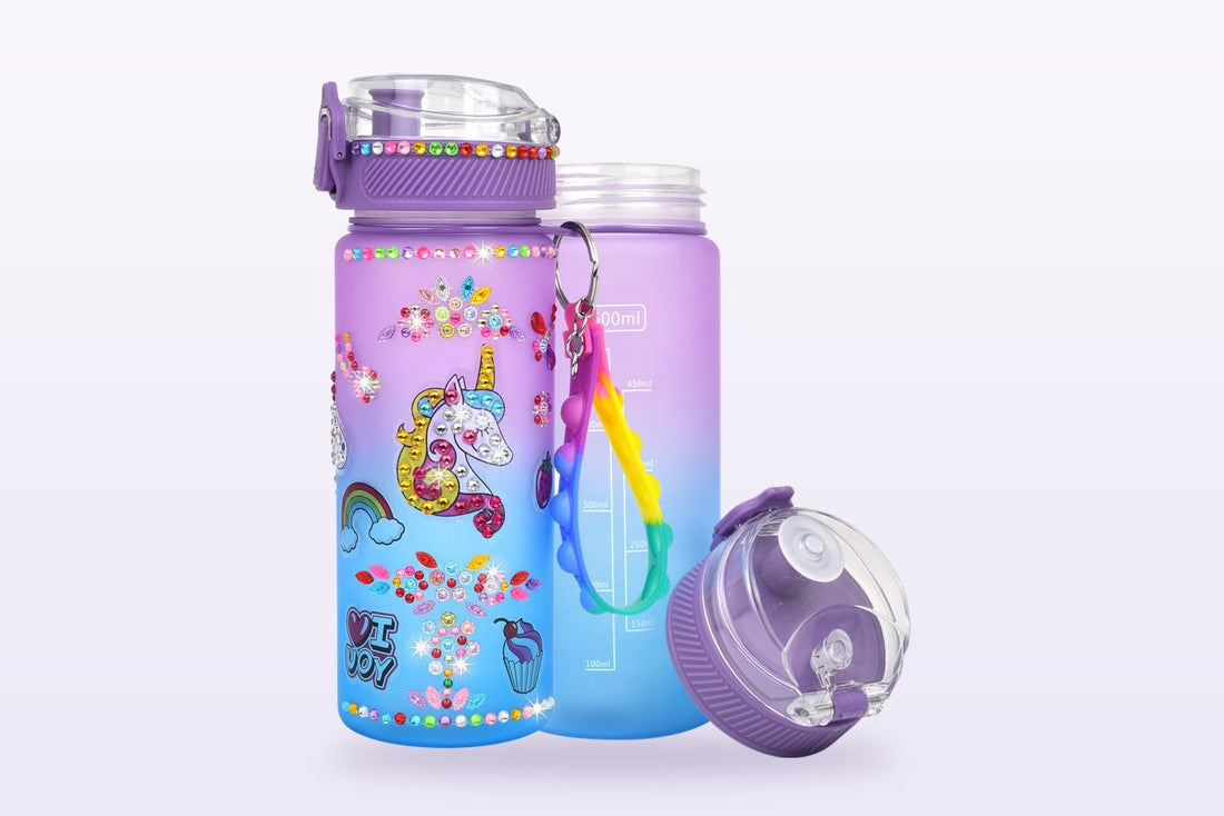 A frosted purple-to-blue water bottle decorated with a gem-covered unicorn, rhinestone flowers, a rainbow and cupcake stickers, beside a plain undecorated bottle and its flip-top cap.