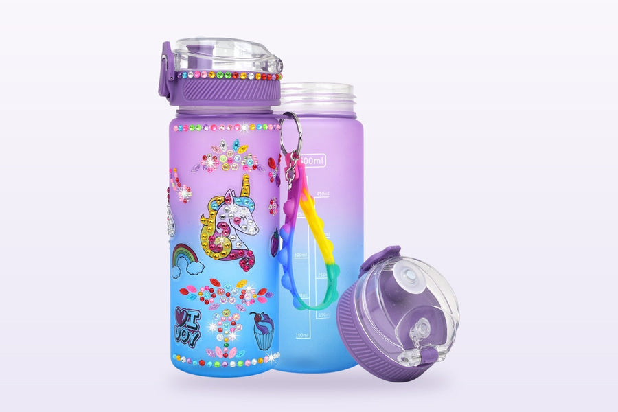 A frosted purple-to-blue water bottle decorated with a gem-covered unicorn, rhinestone flowers, a rainbow and cupcake stickers, beside a plain undecorated bottle and its flip-top cap.