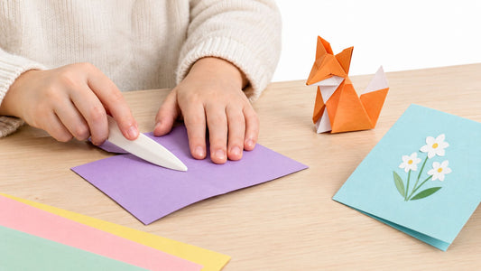 A child's hands pressing the fold of a purple paper card flat with a white bone folder on a wooden table, beside an orange origami fox, a folded blue card with white flowers and sheets of colored paper.