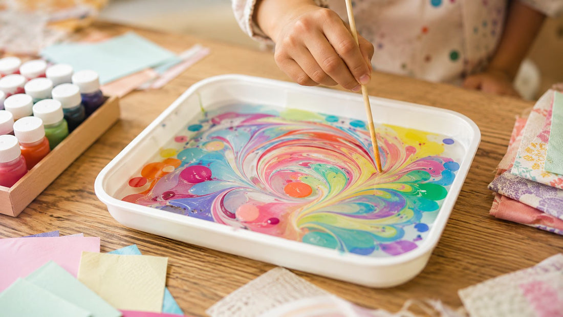 A child dragging a wooden stick through brightly swirled paint floating in a white tray, with paint bottles and marbled paper nearby.