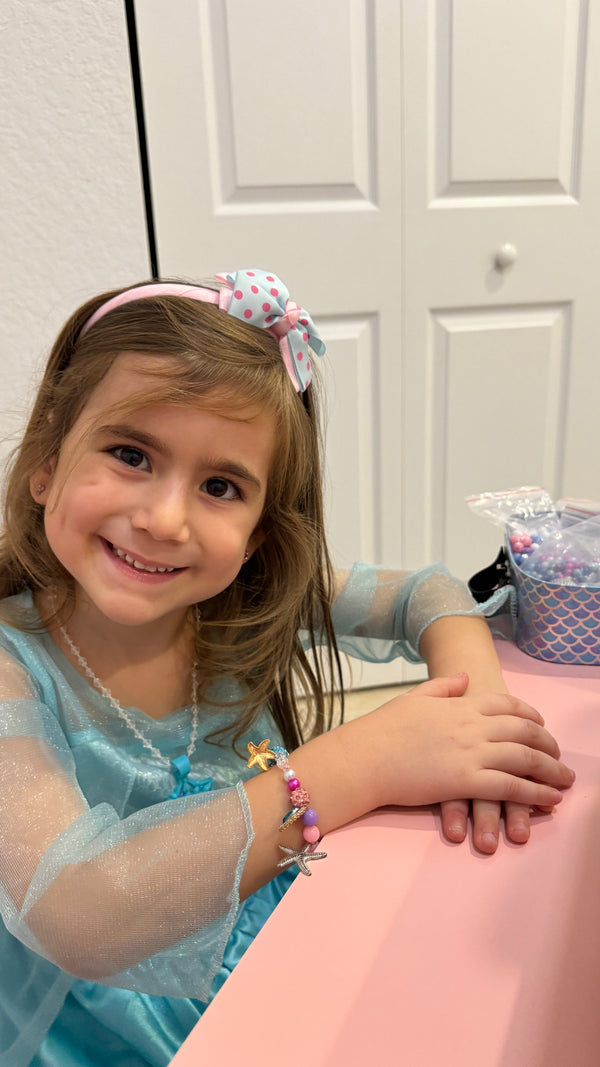 A young girl with brown hair wearing a pink polka-dot bow headband and a light blue dress, smiling while resting her arms on a pink table. She is wearing a colorful beaded bracelet with a sea star charm.