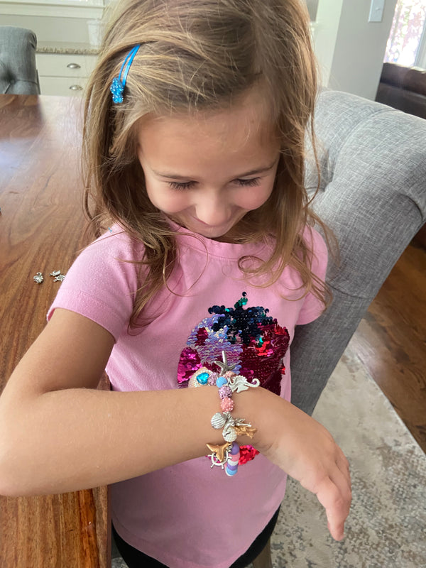 A young girl with light brown hair wearing a pink shirt and blue hair clip looks down at a colorful beaded mermaid bracelet on her wrist, sitting in a gray chair next to a wooden table.
