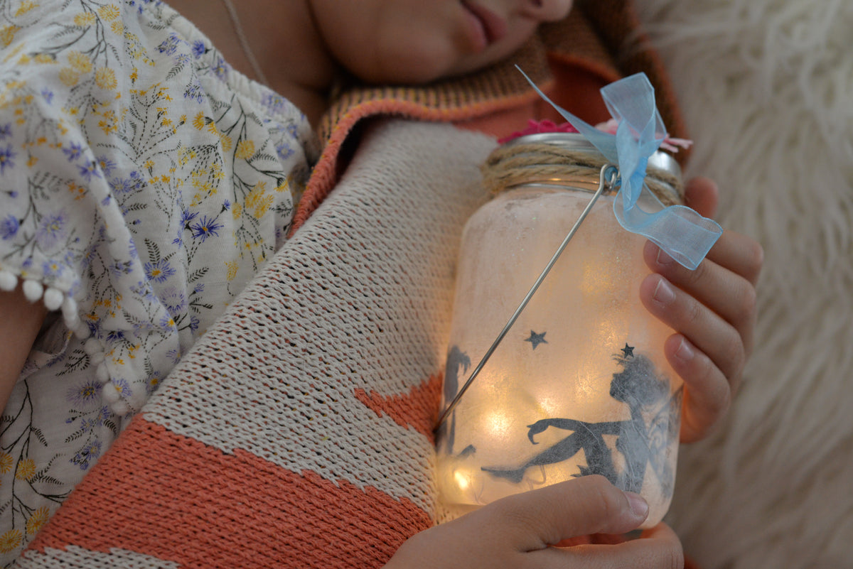 Child holding a finished Pinwheel Crafts Fairy Jar Kit with butterfly and star designs, wearing a floral dress and striped sweater.