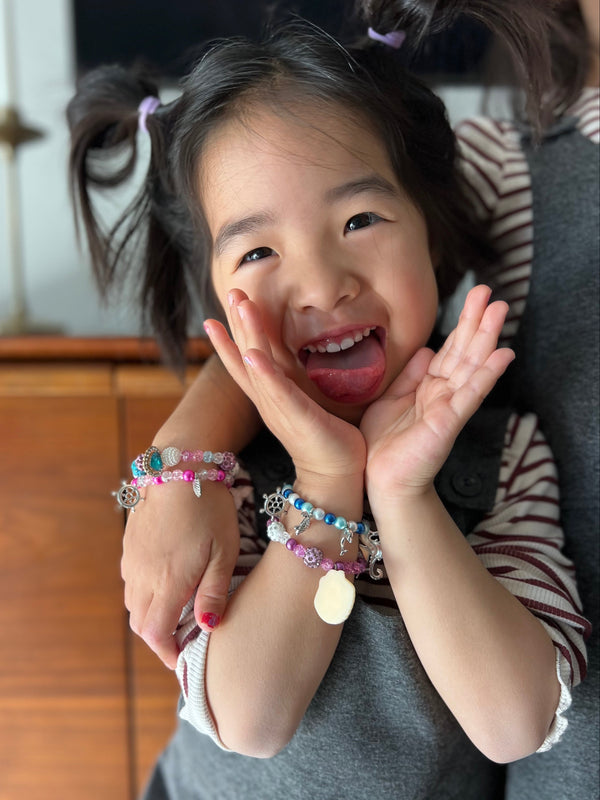 A young child with dark hair in pigtails smiles broadly while wearing multiple colorful mermaid charm bracelets on both wrists. Another person in a striped shirt is partially visible behind them, and the setting appears to be an indoor home environment with a wooden cabinet in the background.