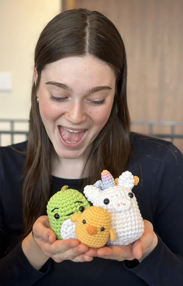 A young woman looks down at the crocheted dinosaur, chick, and unicorn she holds in her hands