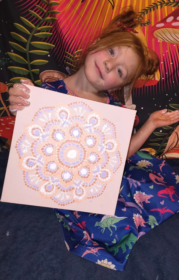 Red-haired girl lying back and smiling as she holds up her finished dotted mandala canvas