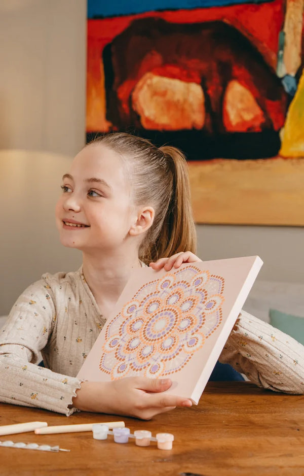 Girl holding up her finished mandala canvas, with paint pots and dotting tools on the table