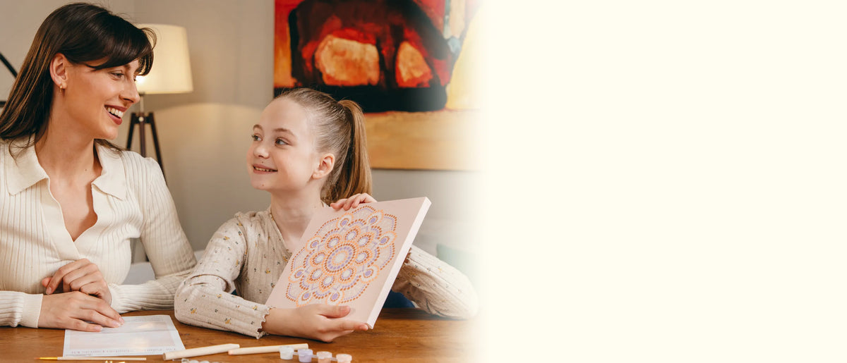 Mother and daughter smiling at each other at the table as the girl holds up her dotted mandala canvas