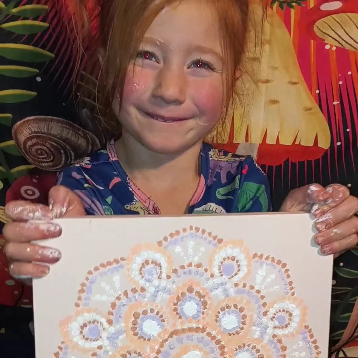 Smiling girl with paint on her hands holding up her finished dot painted mandala canvas