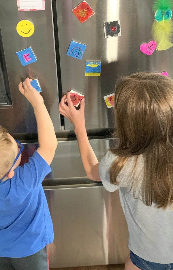 Two kids sticking their painted tile magnets onto the front of a stainless steel fridge