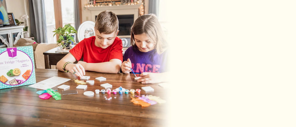 A boy and a girl painting small ceramic tiles at the dining table with the kit box beside them
