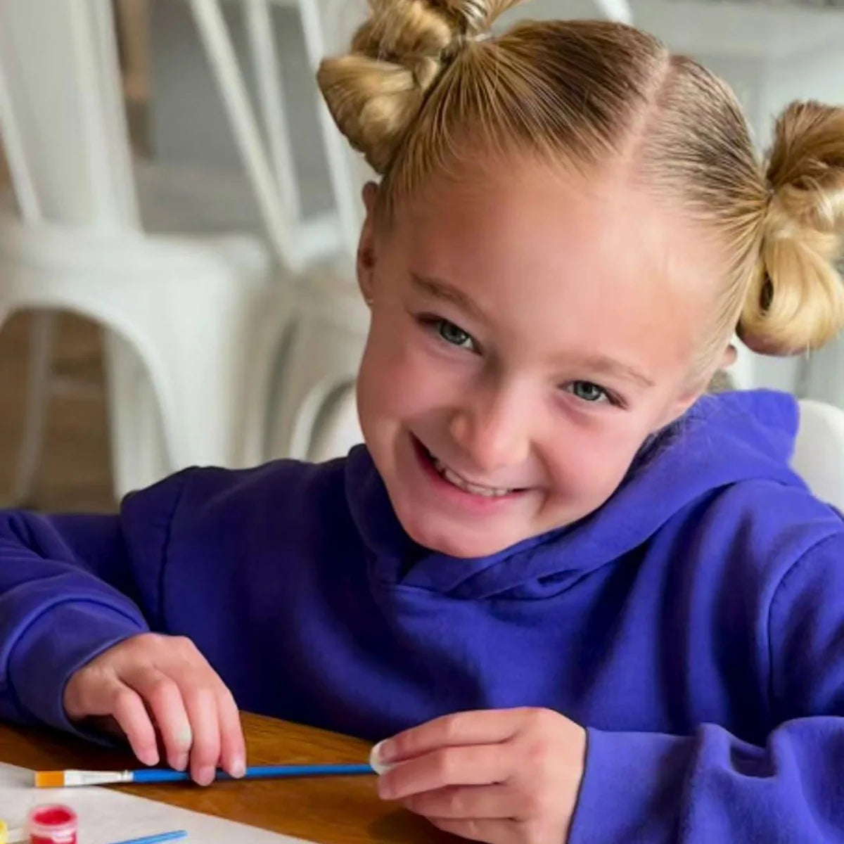 Smiling girl in a purple hoodie holding a paintbrush at the table, ready to paint a tile