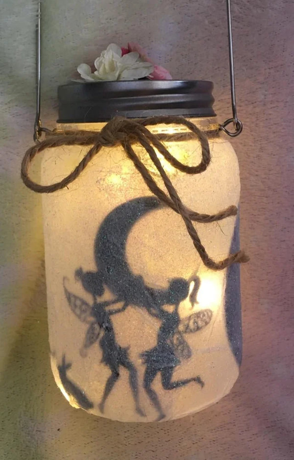 Lit fairy jar with a moon and two fairy silhouettes inside, hanging by its handle with a twine bow