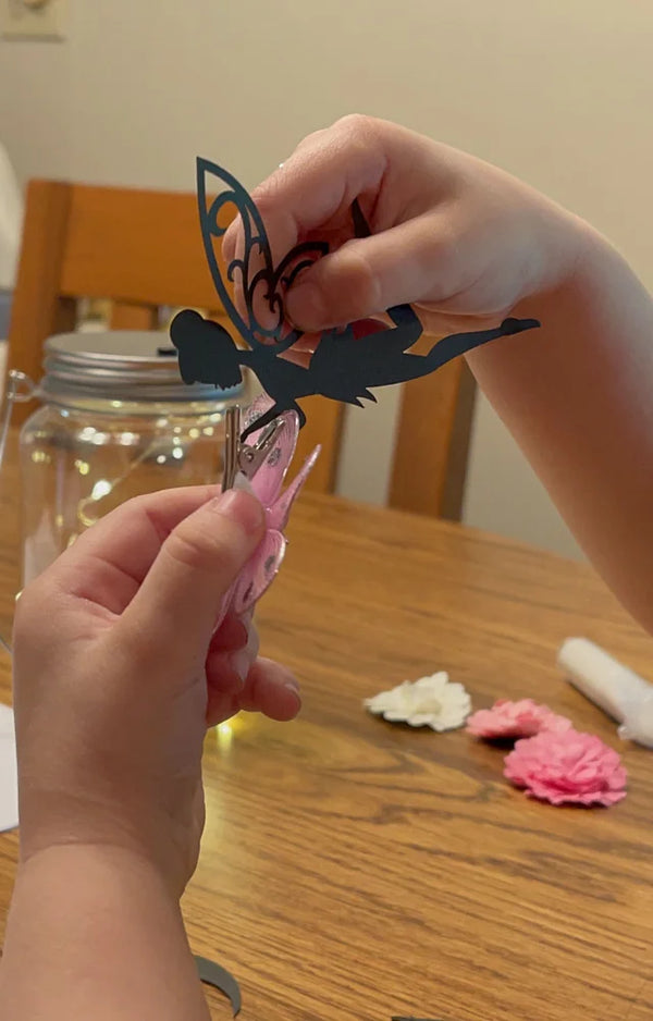 A child's hands holding a fairy silhouette over the jar, with decorative paper flowers on the table