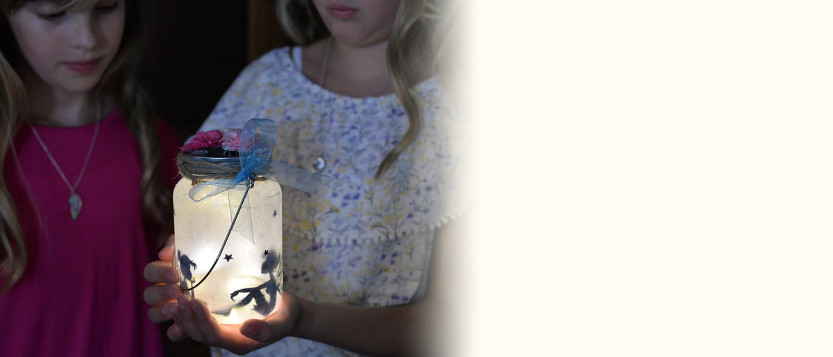Two girls holding a decorated fairy jar in a dim room, its LED lid lighting up the fairy scene inside