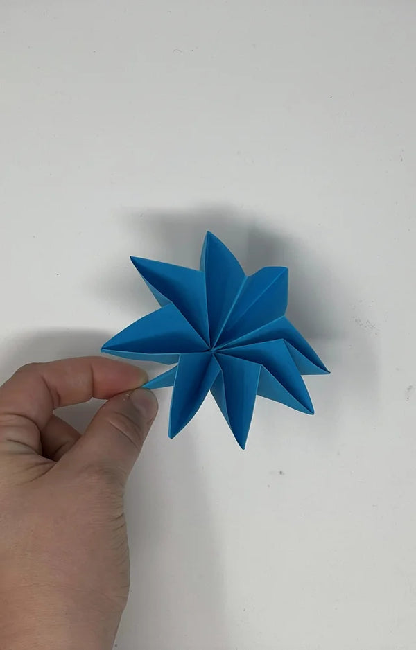 A hand holding a blue star-shaped folded paper flower with pointed petals over a white table
