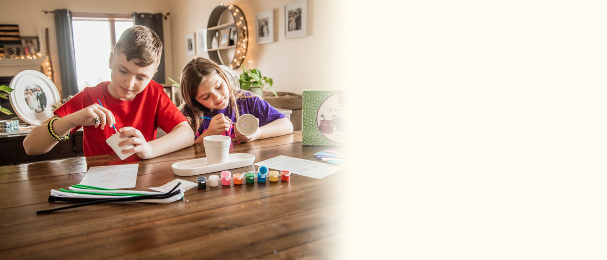 Brother and sister painting flower pots together at the table, paints and brushes in front