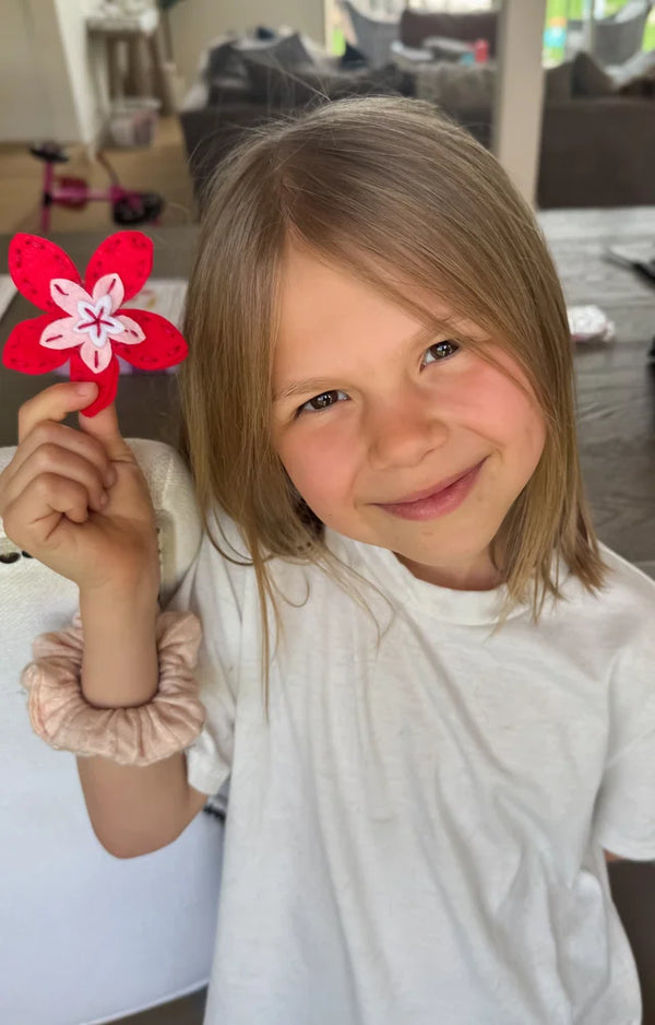 Girl grinning as she holds up the felt flower she sewed from the kit