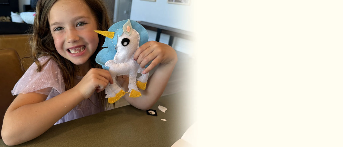 Girl grinning at the table with the felt unicorn she sewed from the Fairytale Sewing Kit