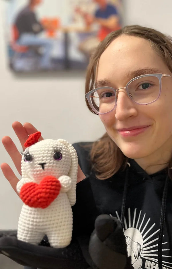 A young woman in glasses holds up her finished crocheted white bunny with a red heart