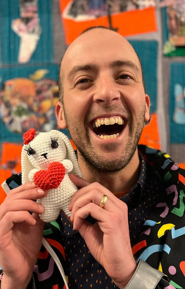 An adult maker laughs as he holds his finished crocheted white bunny with a red heart